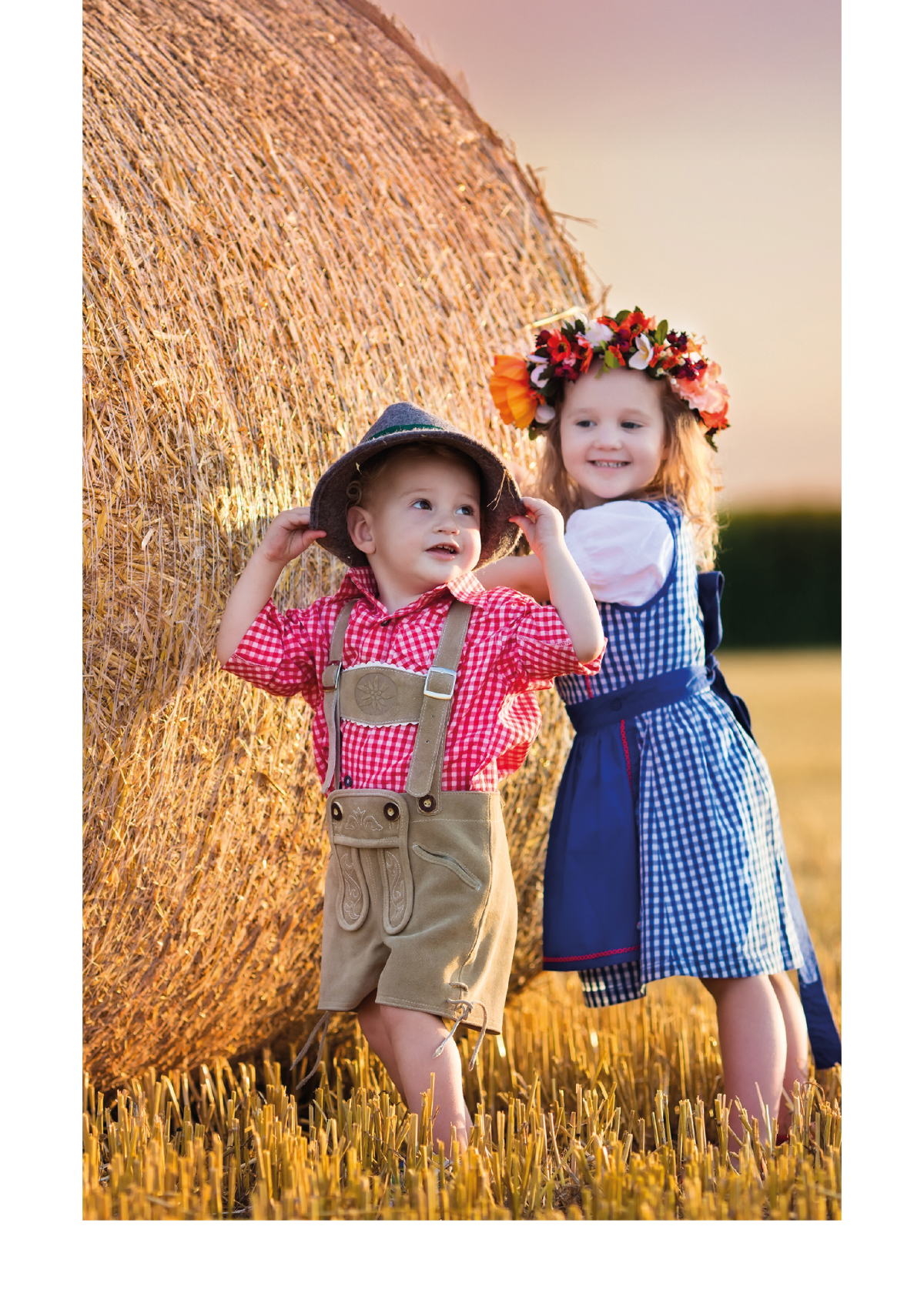 Zwei Kleinkinder in Drindl und Lederhosen vor einem Strohballen.